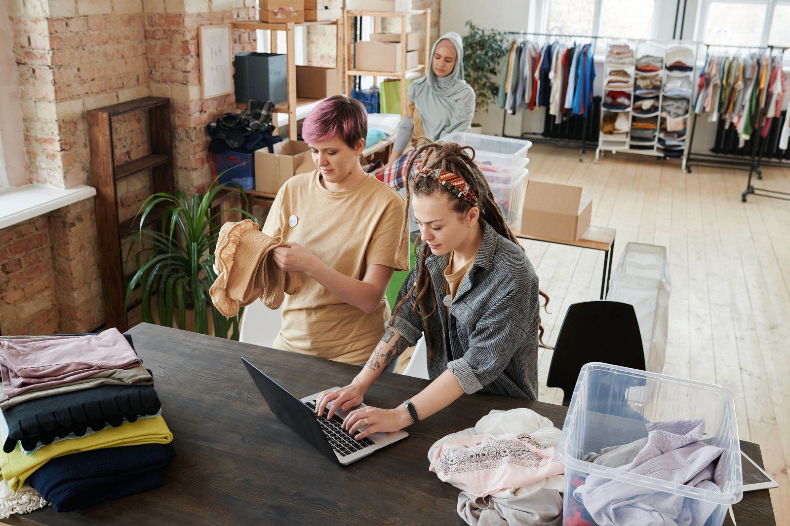 Women organizing clothes and using a laptop in a donation center. Diverse teamwork and collaboration